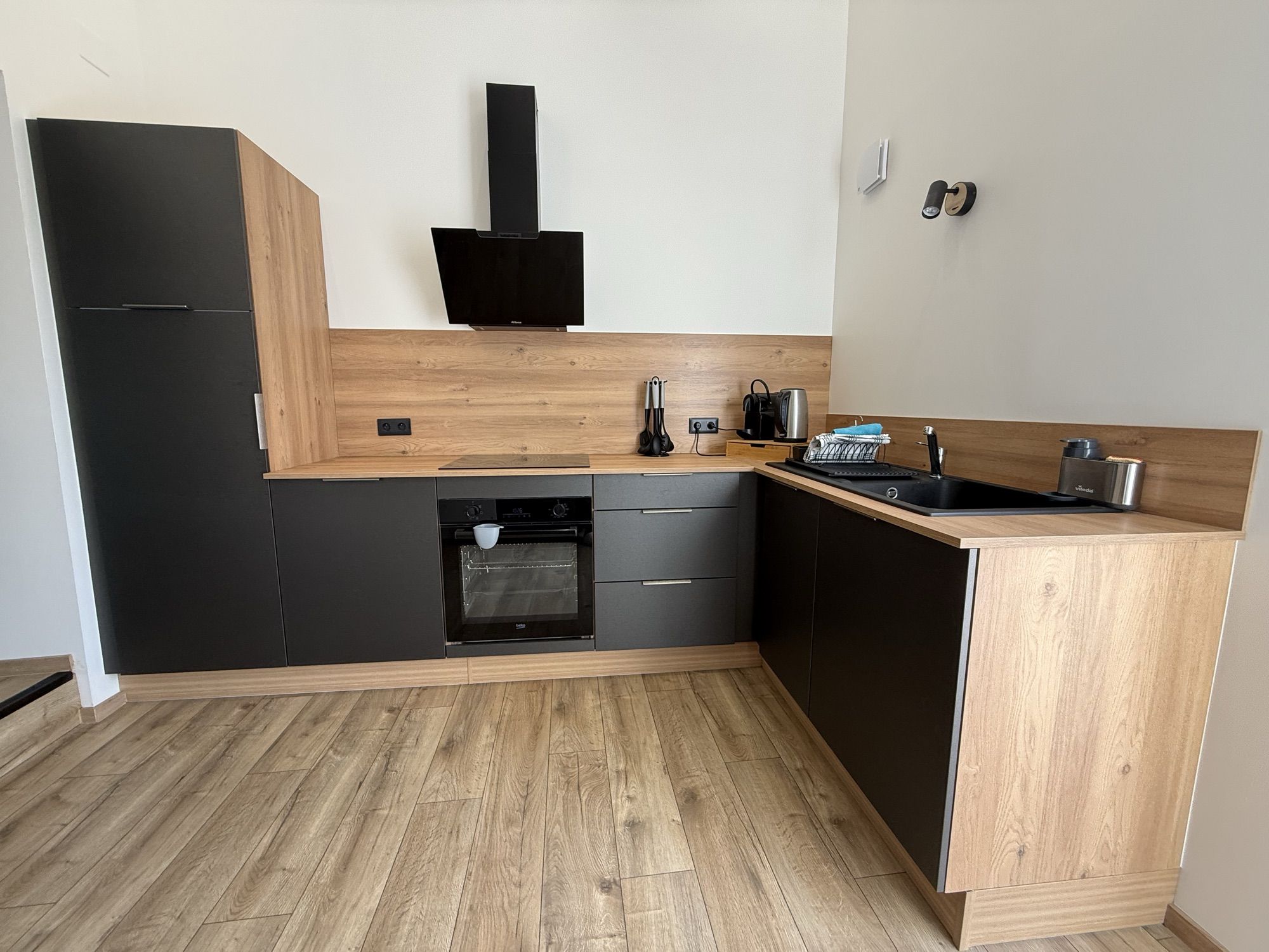 Cuisine en angle, façades noires et plan de travail bois : four, plaques à induction, hotte, évier